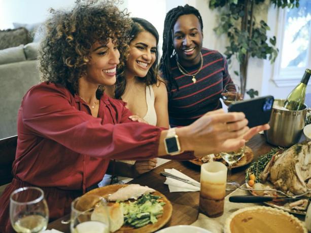 kolm naist teevad õhtusöögilaua kohal selfiet, ämber veiniga, kalkun, kõrvitsapirukas, küünal, taldrikud toiduga ja veiniklaas laual