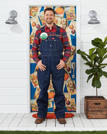 mees kannab taluniku kostüümi kombinesooniga, ruuduline särk, usda orgaaniline nööp seisab halloweeniks kaunistatud ukse ees