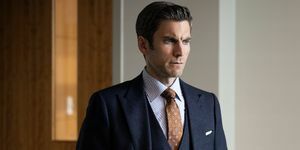 wes bentley tumehallis kolmeosalises ülikonnas pruuni trükitud lipsu ja valge ja sinise triibulise särgiga seisab kontoris, käed taskus