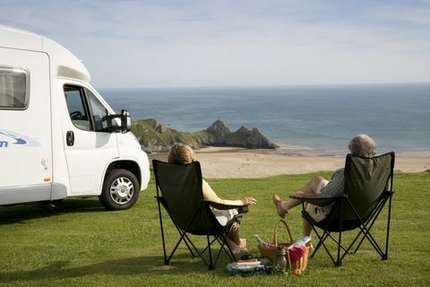 Paar koos piknikuga, mille lõõgastumiseks on toolil oma vagunelamu kõrval nauditav vaade Walesi Goweri poolsaarel asuvale Kolme kalju lahele, Kolmele kalju lahele, Swansea, Wales.