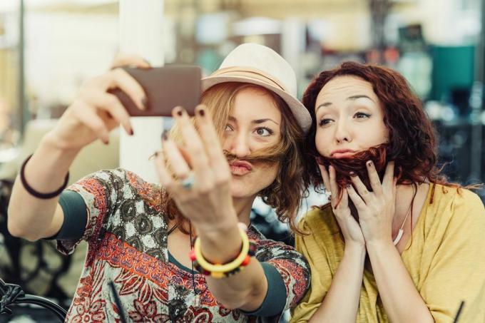 kaks sõbrannat hoiavad juukseid üle näo, et näida välja, nagu neil oleks habe ja vuntsid, ning teevad sellest selfie