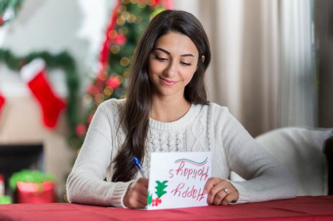 naeratav noor naine istub oma kaunistatud elutoas laua taga ja vaatab keskendunult alla, kui kirjutab sõbrale jõuludeks kirja